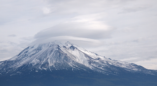 [Photo nearly appears to be black and white but it is in color. There is a whoosh stream cloud sitting right above the top of the mountain. The mountain has lots of snow but there is also rock, trees, and ground visible as darkness up and down throughout many parts of the mountain.]